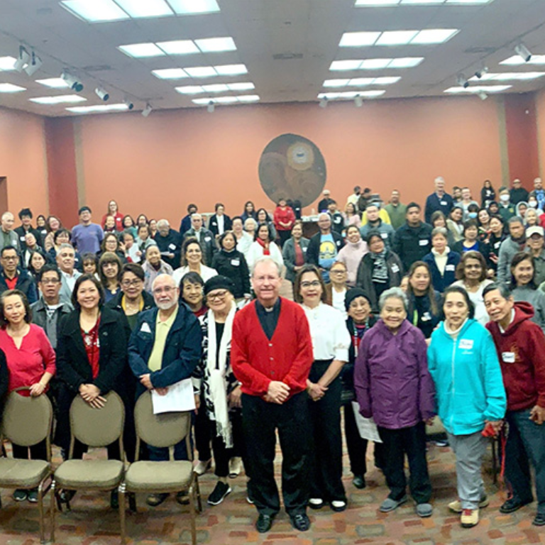 Filipino Community Thumbnail An image of the Filipino Catholic Charismatic community gathered in a meeting hall and listening to a Priest talking on stage.